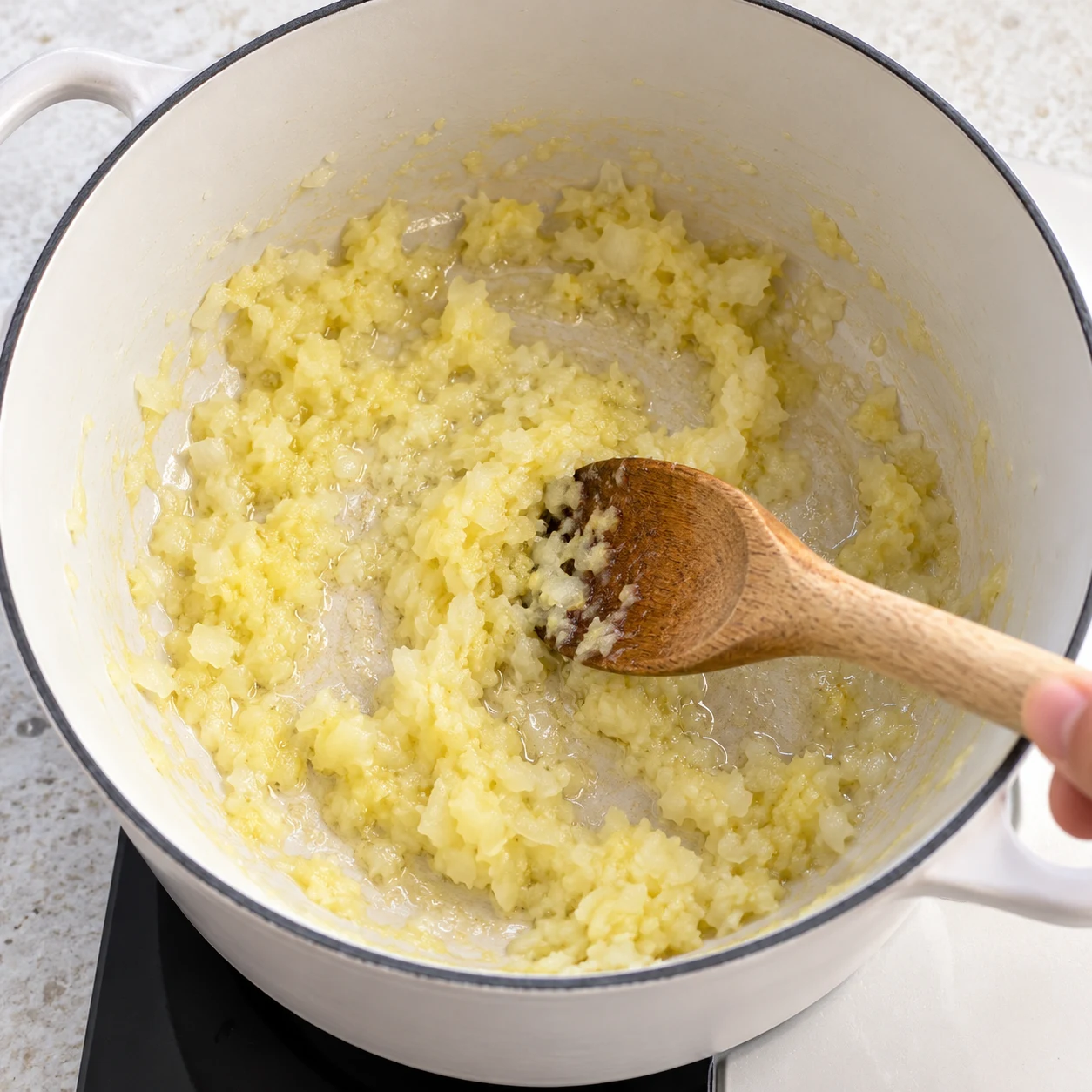 Cuillère en bois remuant une pâte oignon gingembre dans cocotte blanche huilée.