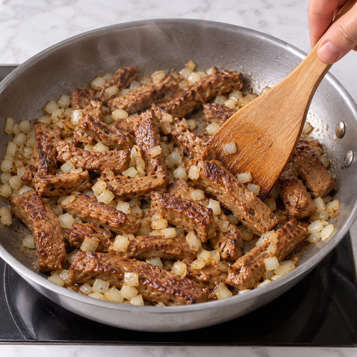 Main anonyme retournant des lamelles de steak végétal avec l’oignon dans la poêle, dorées mais sans sauce, nouilles ni carottes.
