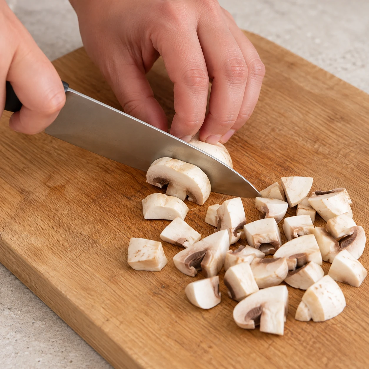 Sur une planche stable, couper un champignon de Paris frais en petits morceaux avec un couteau et une prise en griffe sûre ; aucune sauce ni pâte dans cette scène. Le geste est décrit dans la recette d’origine, non filmé, et la capture source seulement l’état des morceaux.