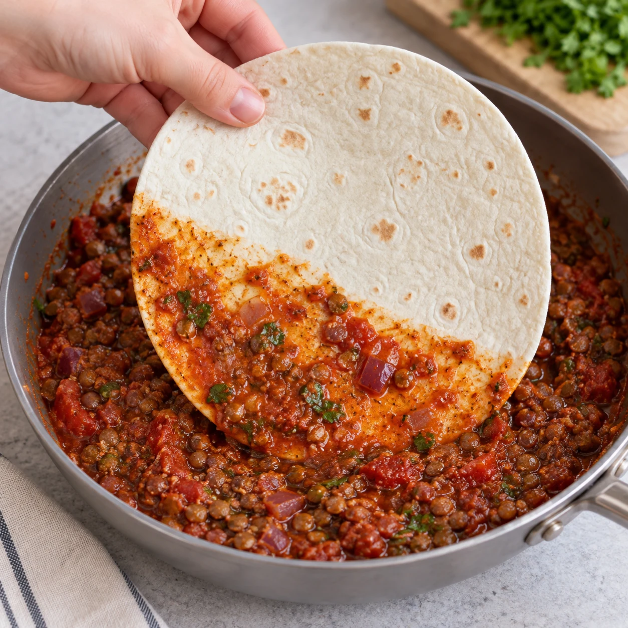 Photographie pédagogique carrée : Trempe une seule face de la tortilla dans la sauce de la garniture, comme dans la recette d’origine. Résultat : Une face porte une fine couche de sauce ; la tortilla reste manipulable. Mains adultes anonymes seulement si nécessaires ; lentilles cuites petites et brunes à la place des haricots rouges de la capture, option source documentée ; fromage végétal au lieu de laitier lorsque présent. Aucun texte.