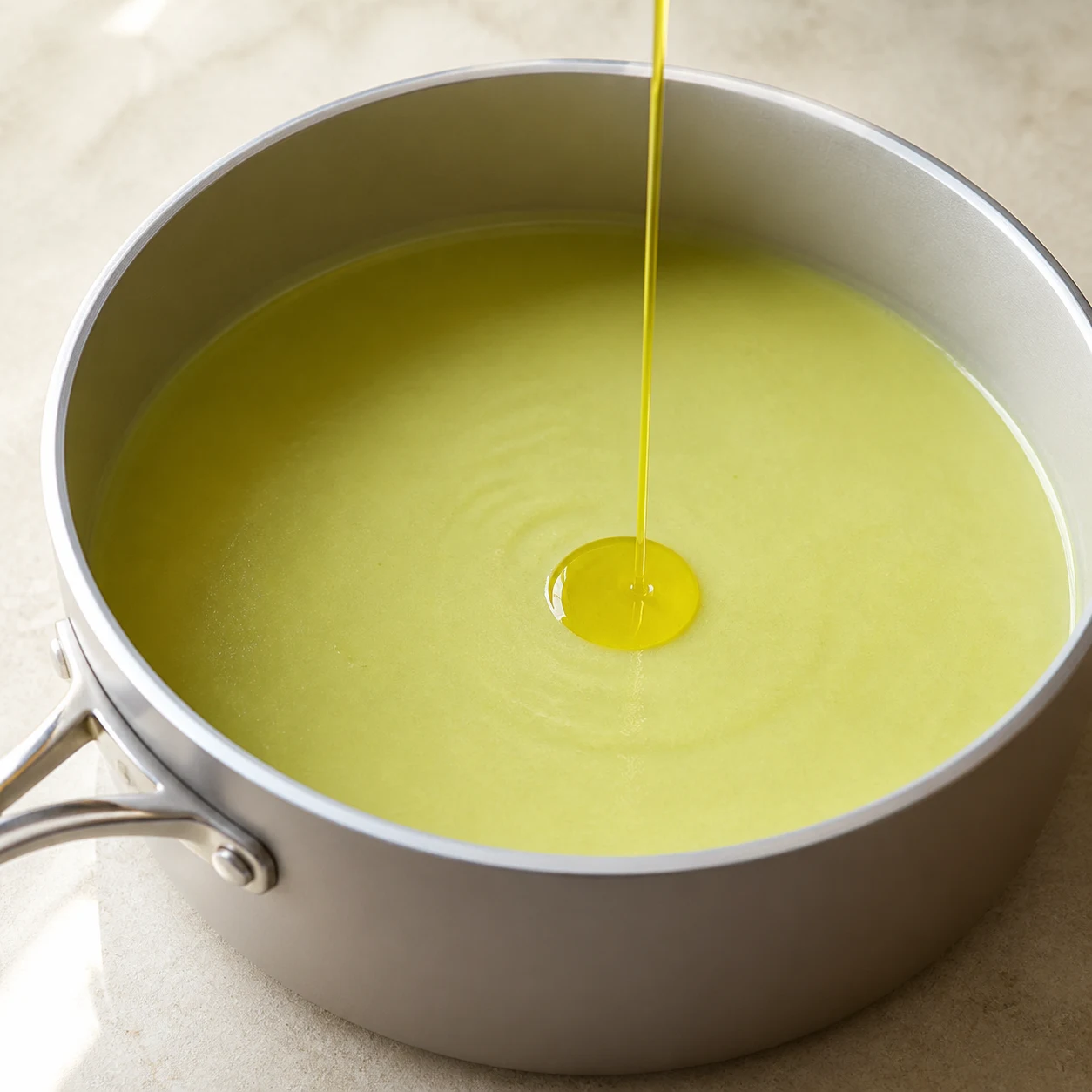 Potage vert pâle tout juste mouliné dans une casserole, filet final d’huile d’olive et cuillère en bois.