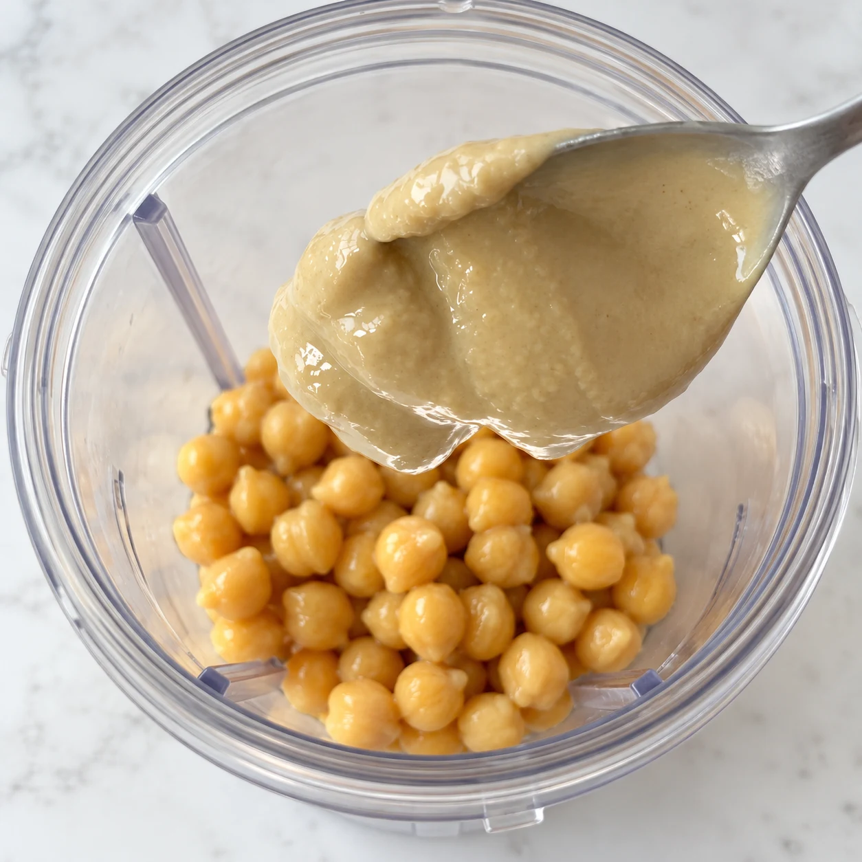 Cuillère de tahini au-dessus des pois chiches dans un bol transparent de mixeur.