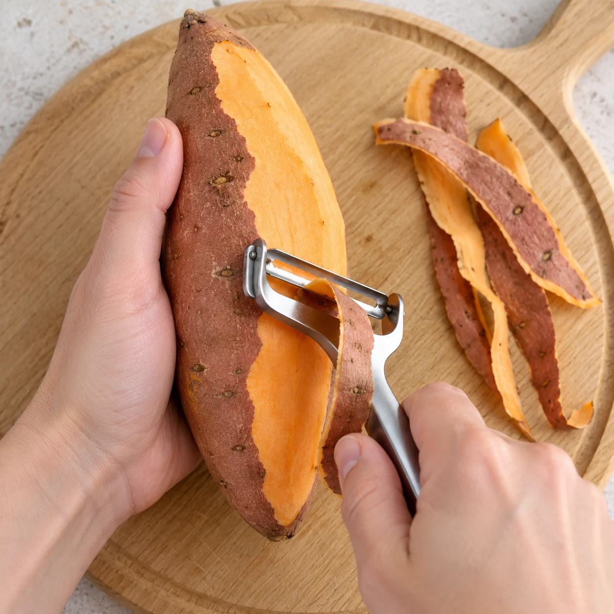Main anonyme tenant une patate douce et économe en action ; chair orange et ruban de peau.