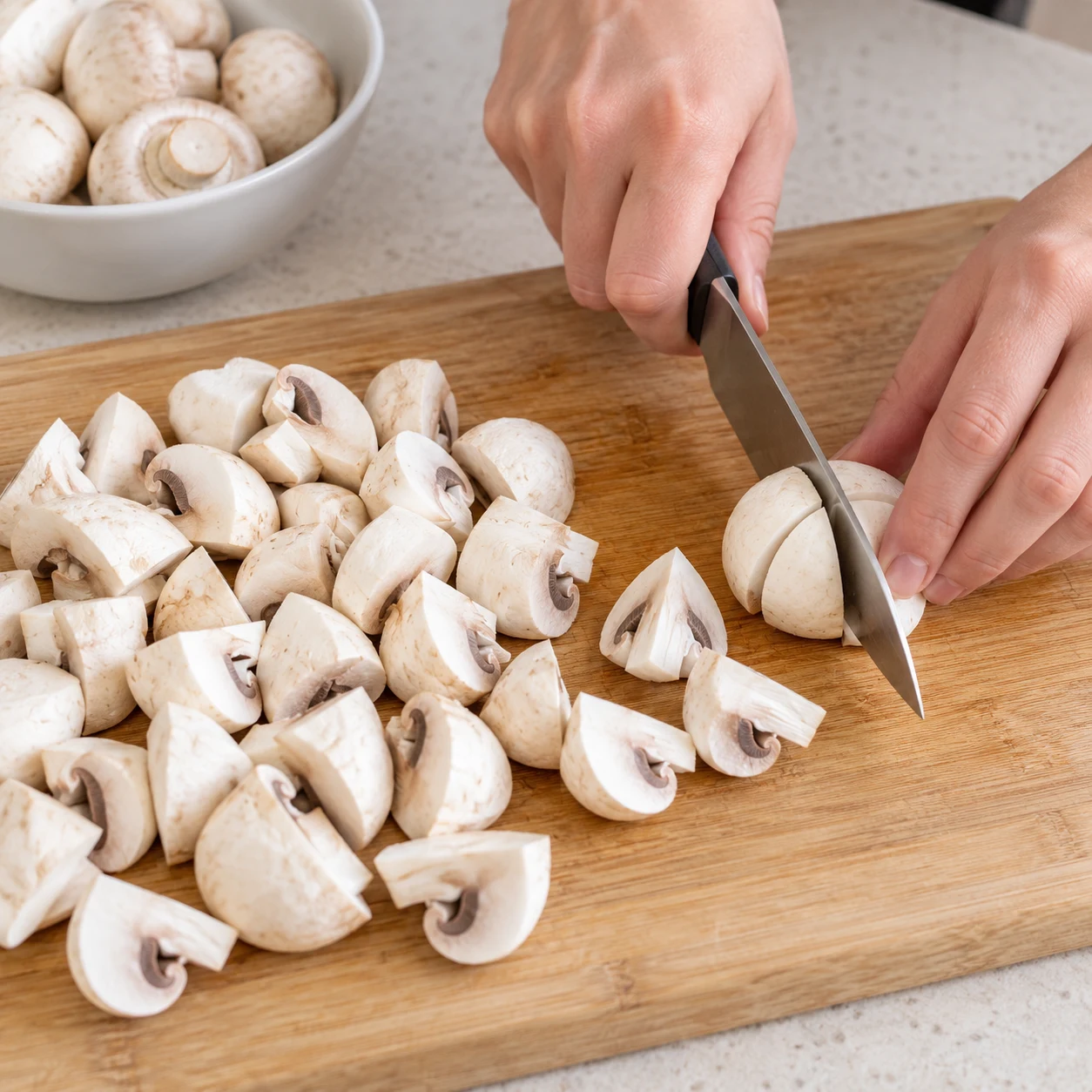 Planche à découper en bois couverte de champignons de Paris blancs coupés en quartiers, une main adulte coupe un champignon au couteau.