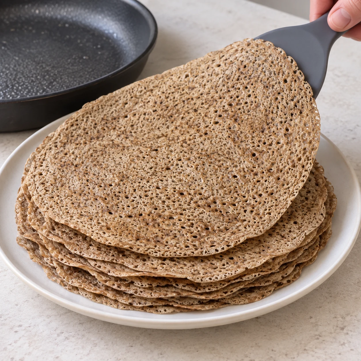 Une galette brune alvéolée est déposée sur une pile de galettes nature, sur une assiette neutre. Aucun jambon, fromage ou œuf.