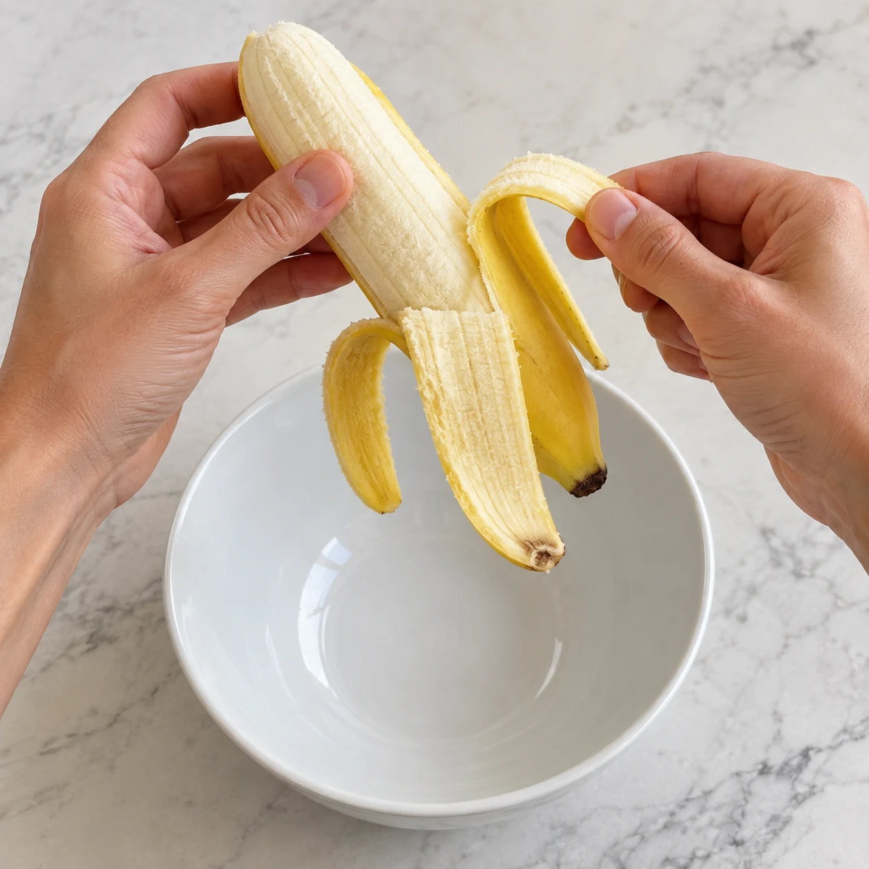 Mains anonymes épluchant une banane au-dessus d’un saladier blanc.