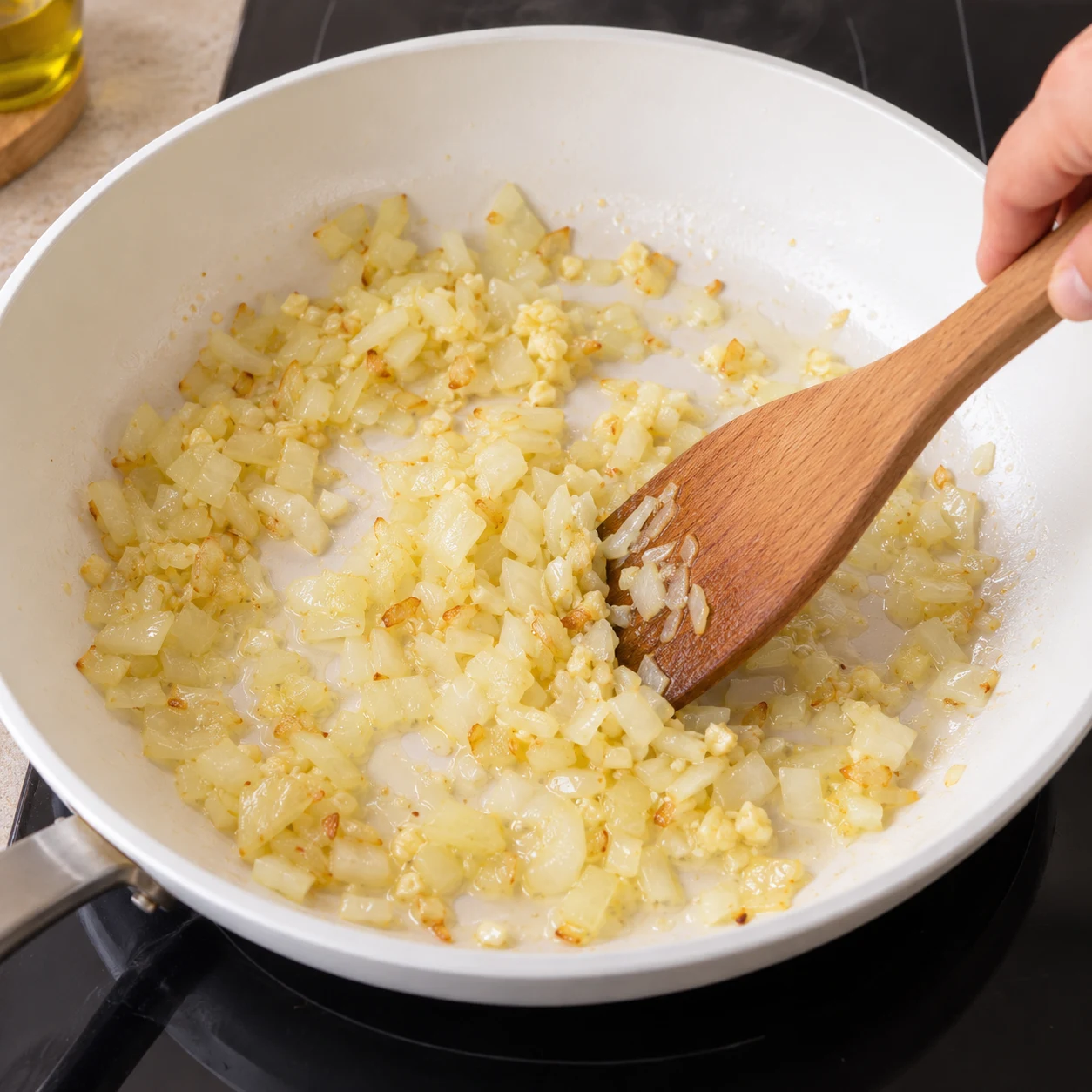 Grande poêle blanche sur une plaque de cuisson, oignon et ail émincés qui revient dans un filet d'huile, spatule en bois.
