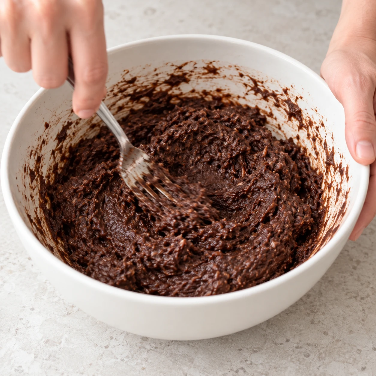Une main anonyme mélange à la fourchette une pâte brun chocolat dense et humide ; aucun œuf, pas de pépites à cette étape.