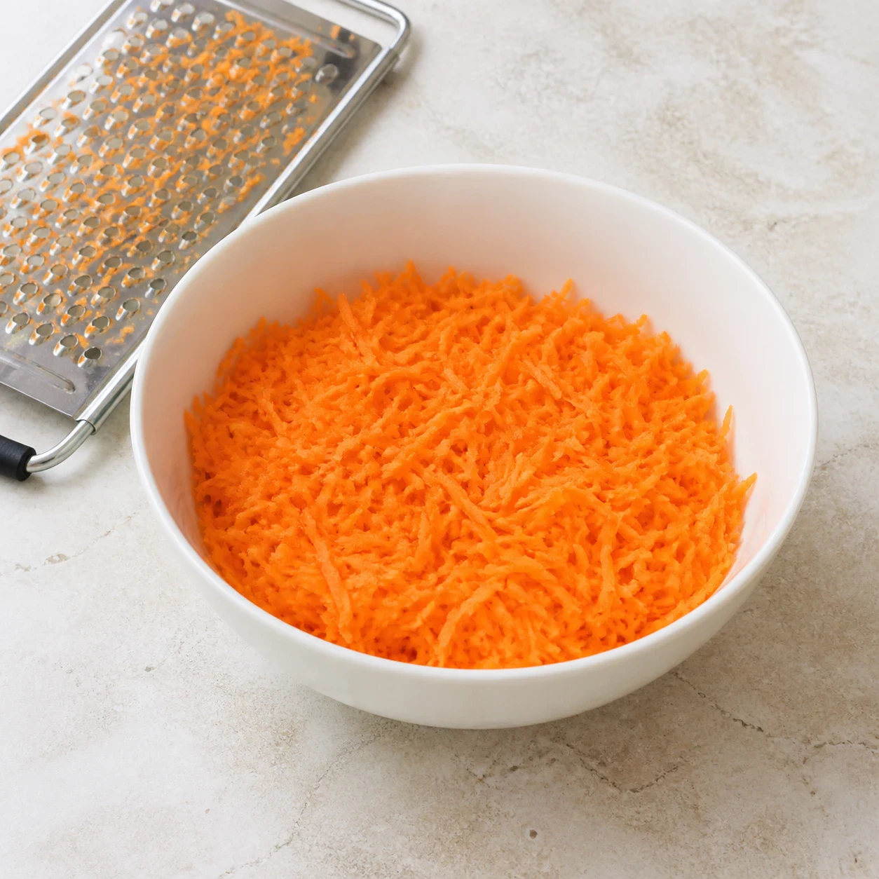 Saladier en céramique blanche rempli de carottes râpées finement, filaments orange vif, râpe posée à côté sur un plan de travail en pierre claire. Aucun autre ingrédient, aucune main, aucun texte.