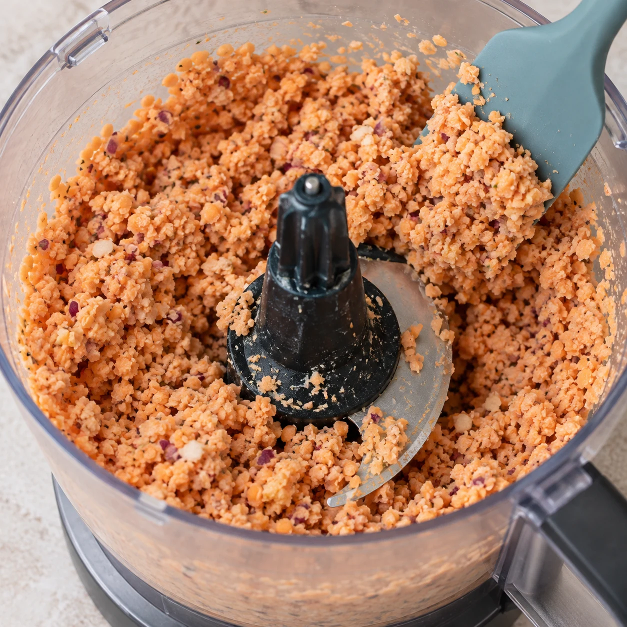 Mixer la base : Mixez les ingrédients dans le robot à lame en S jusqu’à obtenir une pâte granuleuse, comme dans la recette d’origine. Résultat visible : Les morceaux d’oignon et d’ail sont incorporés à une pâte granuleuse de lentilles. Une seule photo carrée de cette phase exacte ; aucun dip, garniture ou ingrédient futur.