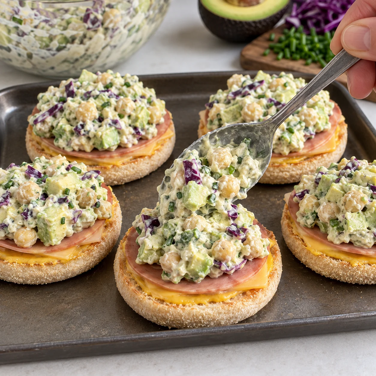 Cuillère déposant la farce épaisse aux pois chiches, avocat, chou rouge, ciboulette et yaourt sur un muffin ouvert déjà garni de tranches végétales et moutarde.