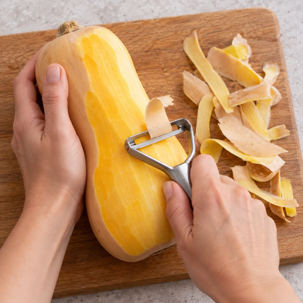 Une main anonyme épluche une butternut sur une planche stable.