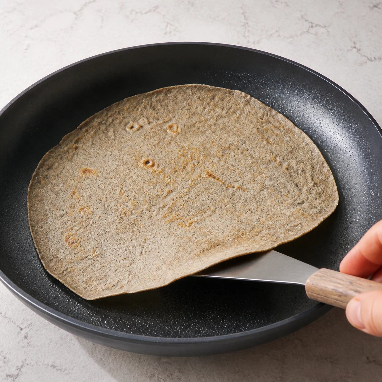 Galette de sarrasin fine en cuisson sur une grande poêle noire légèrement graissée, bords qui se décollent.