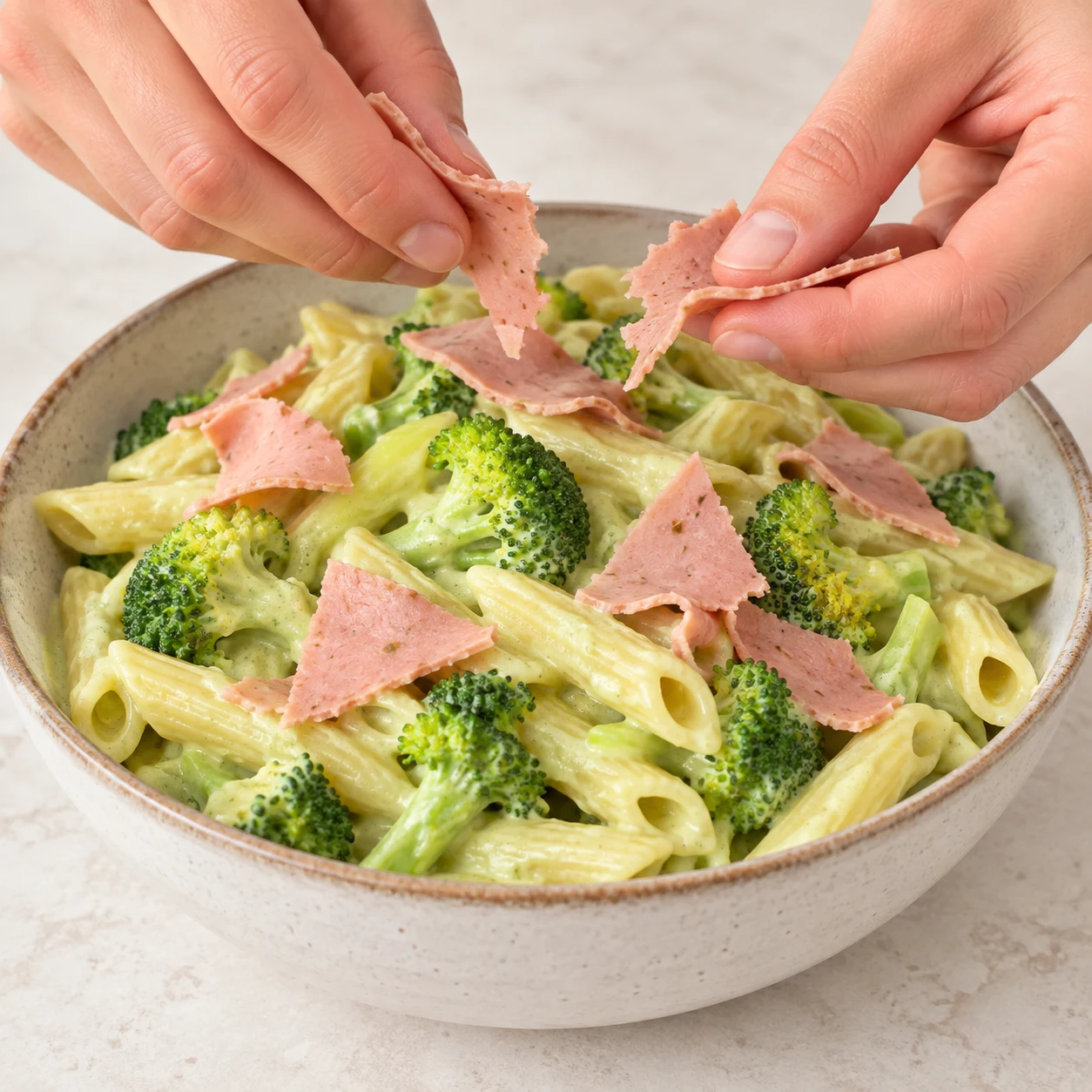 Une main adulte anonyme dépose quelques morceaux de fines tranches roses de jambon végétal aux pois et fèves sur les penne au brocoli en sauce vert pâle. Pas de tofu, jambon animal ni pignons à ce stade.