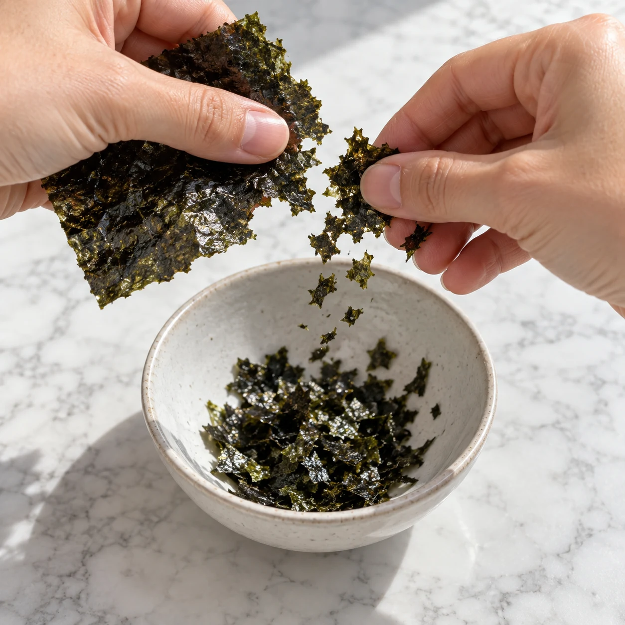 Mains anonymes émiettant une feuille noire verte de nori au-dessus d’un petit bol.