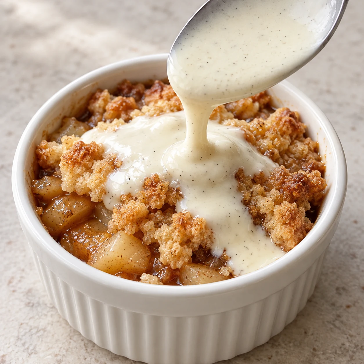 Une cuillère verse une sauce de crème de soja vanillée fluide sur un crumble pomme-poire doré ; pas de chantilly ni de pics.