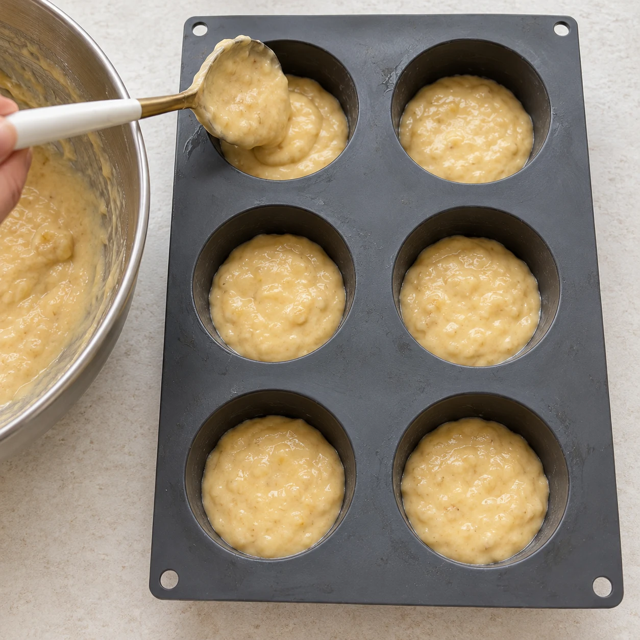Une cuillère remplit les six empreintes grises de pâte à la banane.