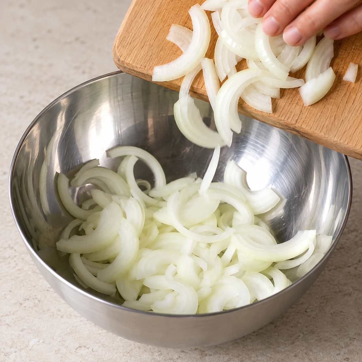 Lamelles blanches d’oignon glissant d’une planche dans un saladier en inox ; mains anonymes, aucun autre ingrédient.
