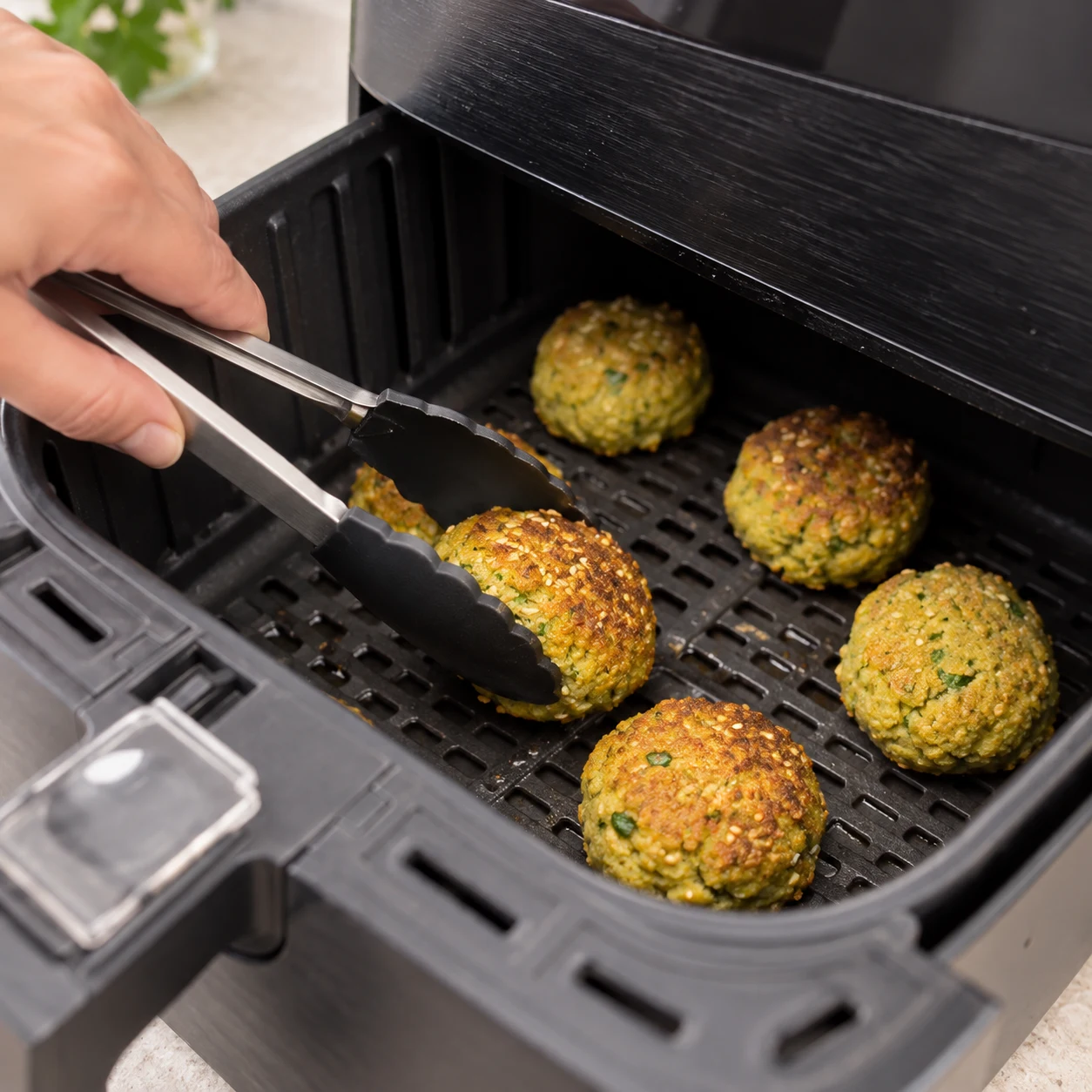 Une main anonyme avec une pince retourne un falafel dans le panier ouvert ; les falafels sont partiellement dorés à mi-cuisson.