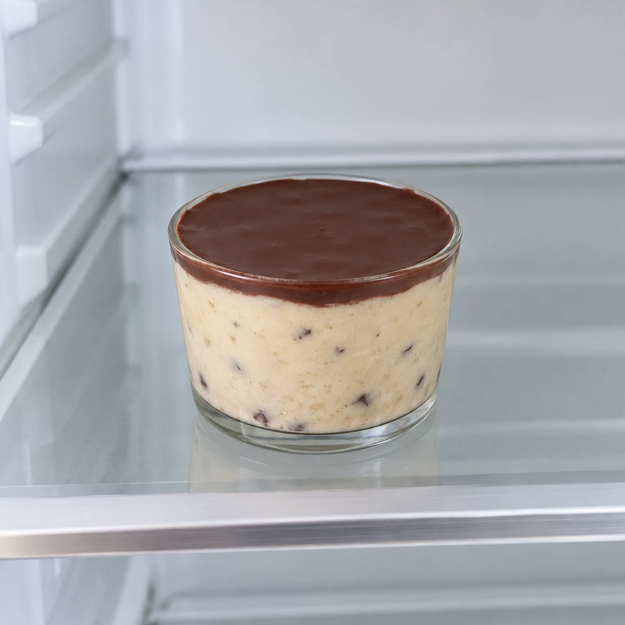 Petit verre de pâte beige à pépites couvert d’une fine coque de chocolat dans un réfrigérateur.