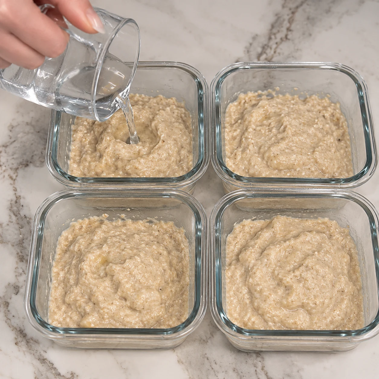 Main versant un petit filet d’eau dans une pâte beige de bowl cake en verre, pas de poudre chocolatée.