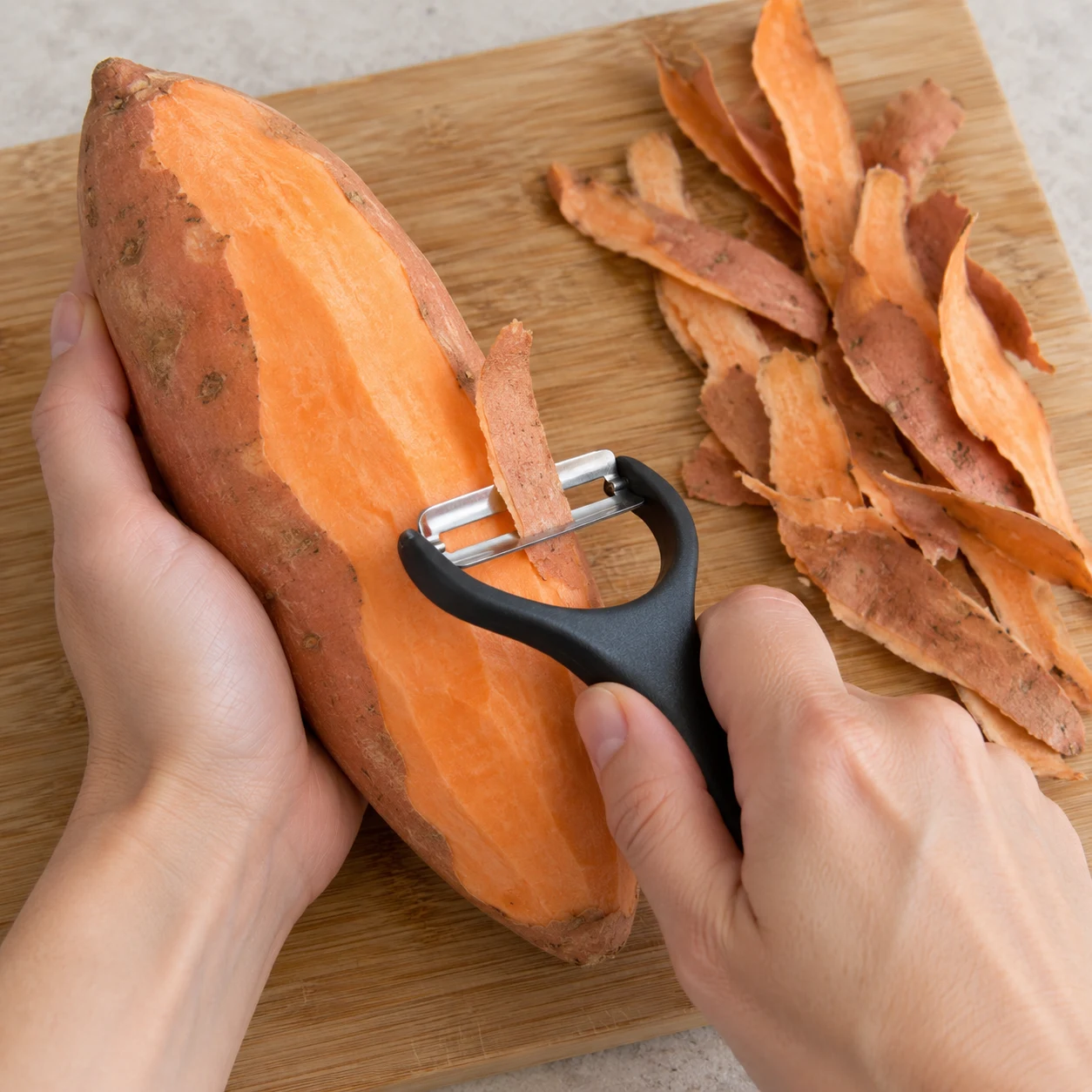 Main anonyme avec économe épluchant la patate douce sur une planche.