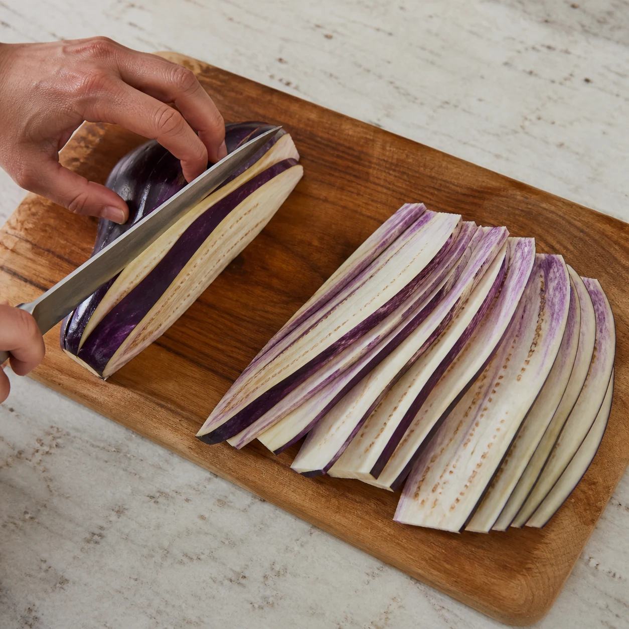 Main adulte en prise griffe découpant une aubergine pelée en fines lamelles sur planche stable.