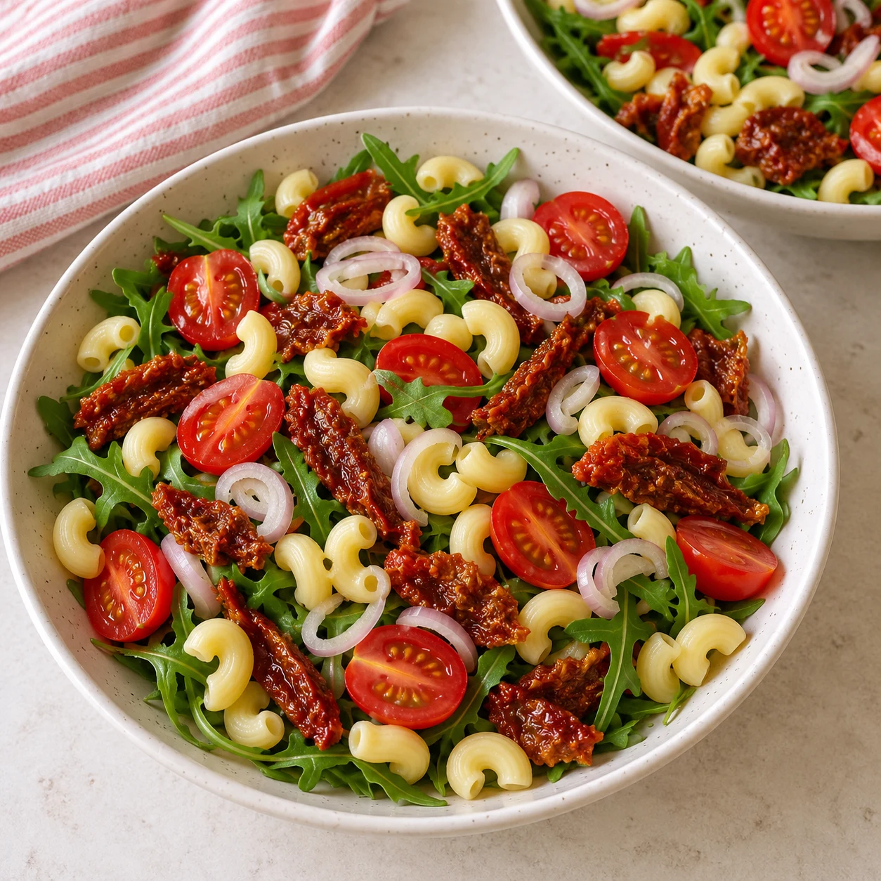 Salade en cours de montage avec uniquement roquette,pâtes,tomates cerises,tomates confites et fines rondelles échalote. Les pâtes éventuelles de cette adaptation sont des coquillettes courtes jaunes courbées et creuses, pas des radiateurs.