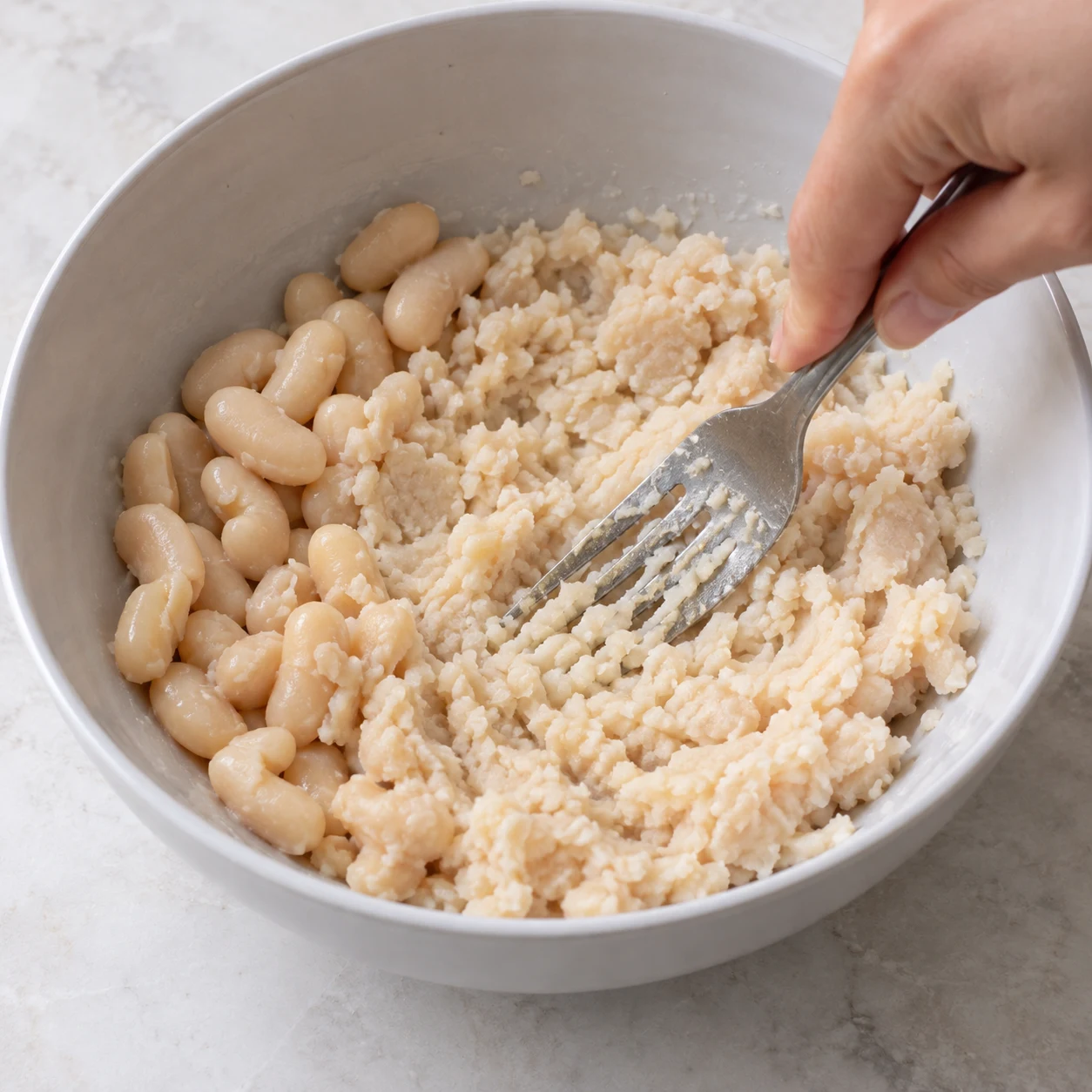 Fourchette écrasant des haricots blancs cuits égouttés dans un saladier neutre, petits grains et purée claire.
