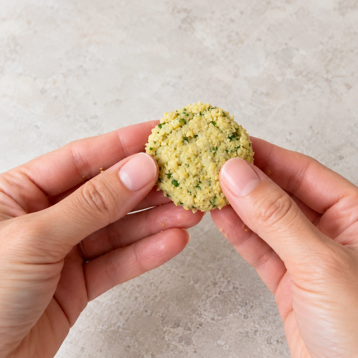 Deux mains adultes anonymes façonnant un petit palet jaune pâle de falafel cru avec points verts.
