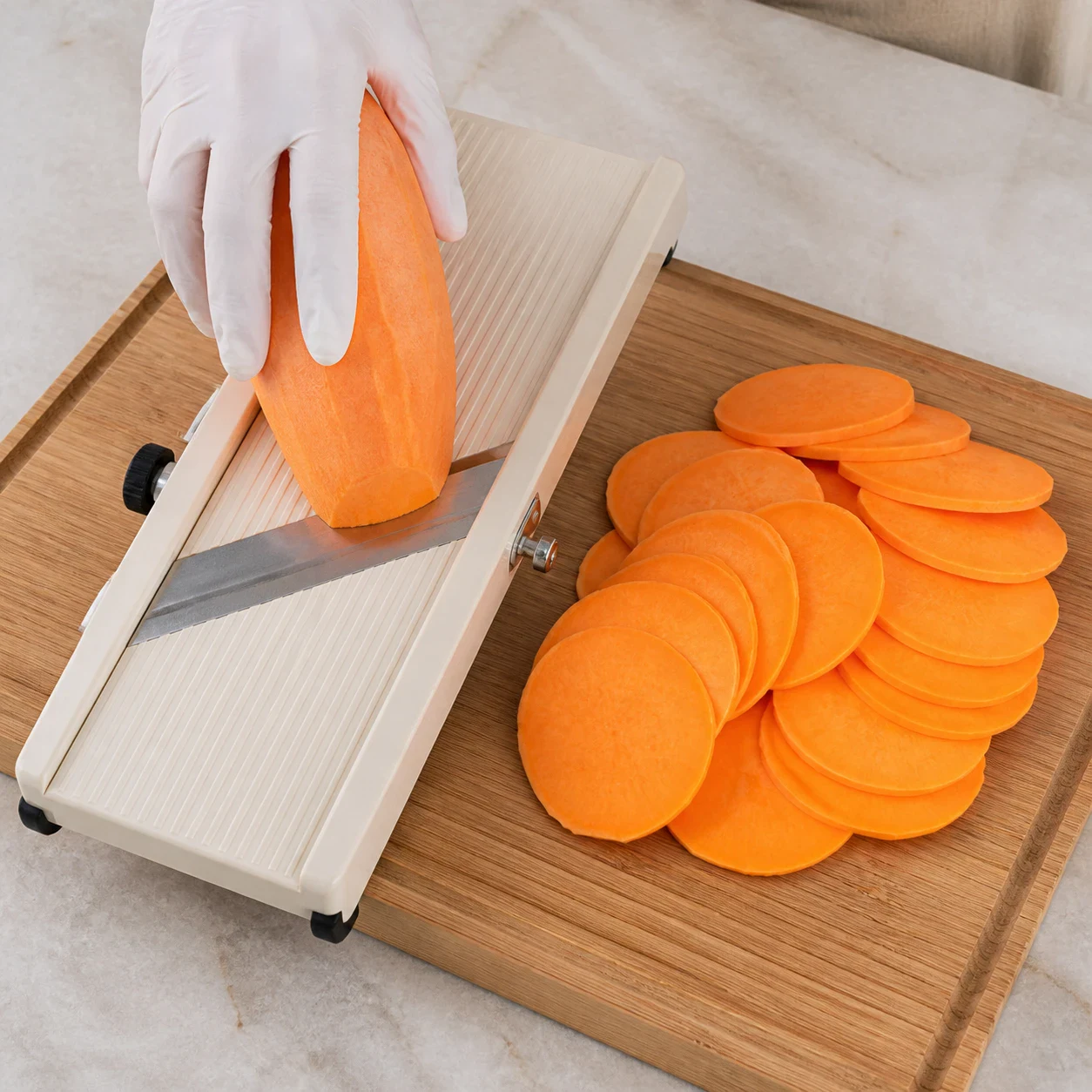 Patate douce épluchée et tranches de patate douce orange de deux millimètres d'épaisseur étalées à côté d'une mandoline de cuisine, une main d'adulte gantée tenant le légume, aucun autre ingrédient, aucun texte, aucune marque.