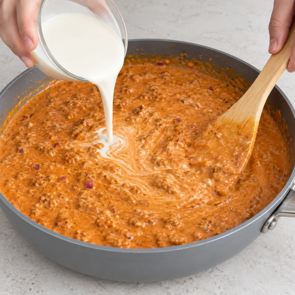 Ajoute les 150 g de crème de soja cuisine et mélange pour obtenir une sauce homogène. Montrer seulement les ingrédients de cette phase ; mains anonymes, sauteuse grise et plan de travail pierre pâle.