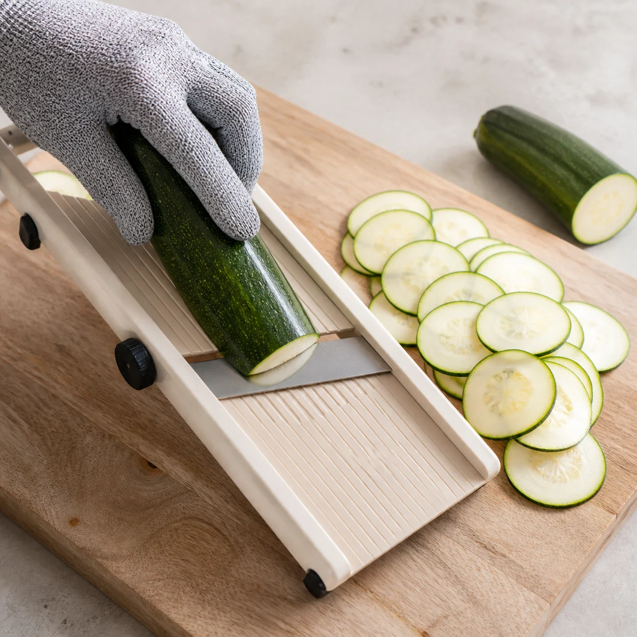 Courgette entière et rondelles de courgette de deux millimètres d'épaisseur posées à côté d'une mandoline de cuisine, une main d'adulte gantée maintenant le légume au-dessus de la lame, aucun autre ingrédient, aucun texte, aucune marque.