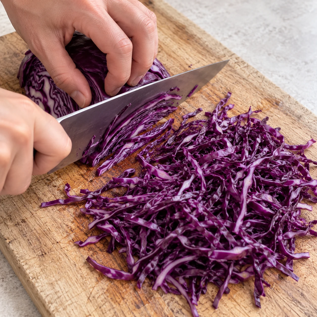 Planche en bois avec un quart de chou rouge en partie émincé en filaments fins violets, couteau de chef et mains adultes anonymes en position de griffe.