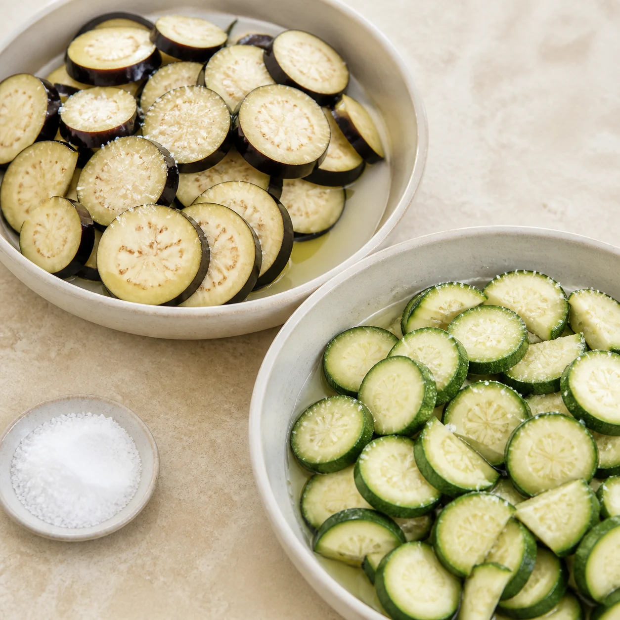 Deux plats séparés d’aubergines et de courgettes en rondelles légèrement salées, avec eau de végétation visible.