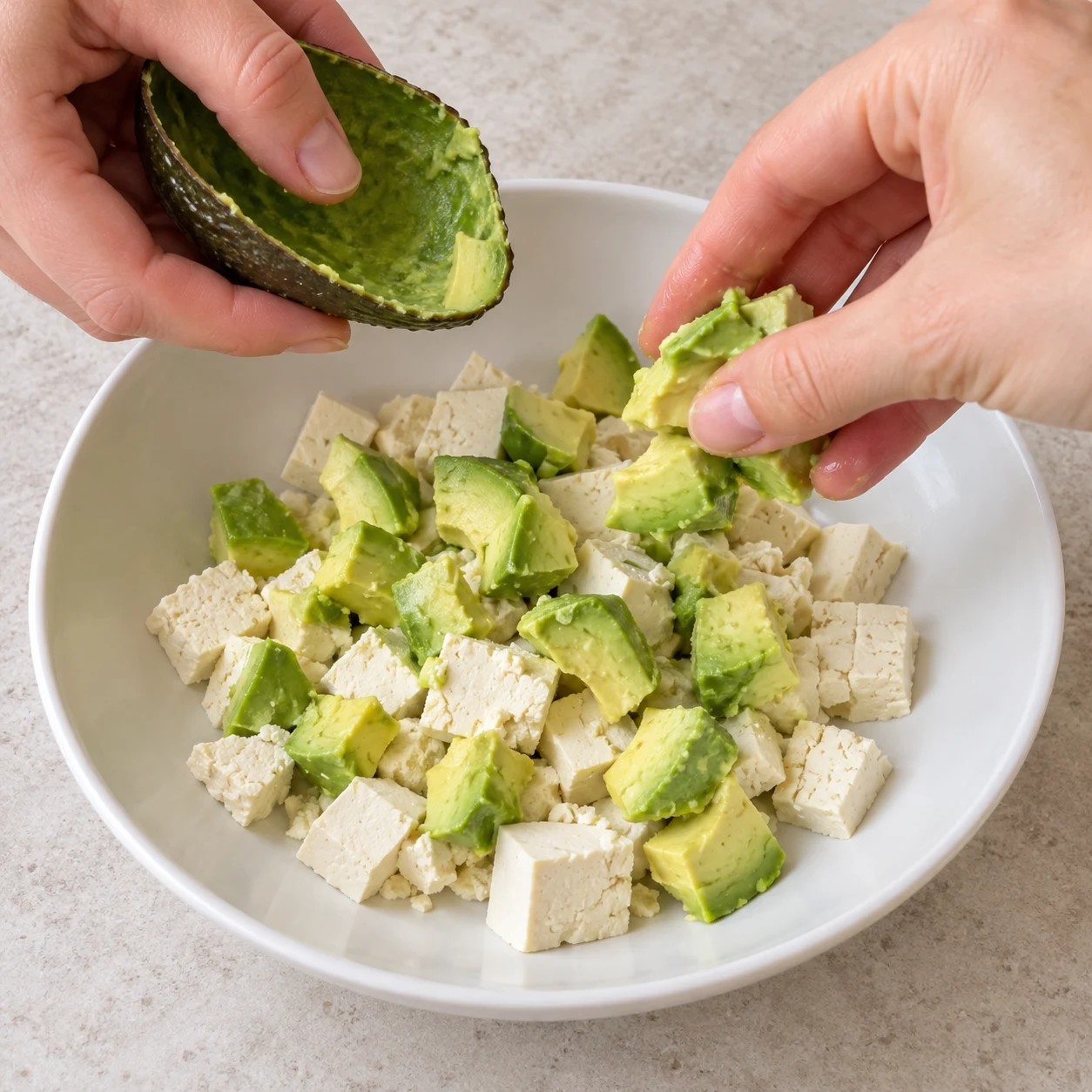 Deux mains adultes anonymes retirent la peau du demi-avocat et déposent des morceaux de chair dans une assiette de petits morceaux blancs de tofu nature stérilisé. Aucun couteau ni œuf.