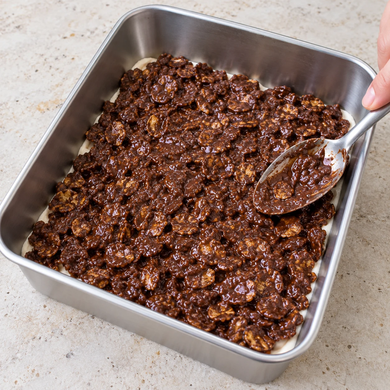 Moule en inox avec la couche de crème claire au fond, recouverte de morceaux de mélange chocolat noir et corn flakes enrobés répartis à la cuillère.