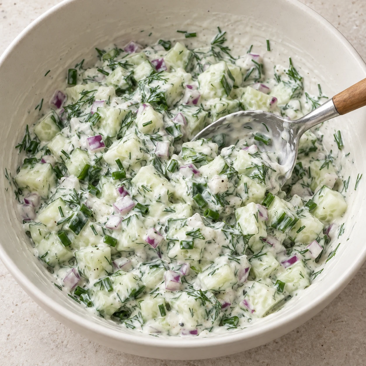 Cuillère mélangeant la sauce blanche soja-moutarde aux petits cubes de concombre pâle, oignon rouge, aneth et ciboulette.