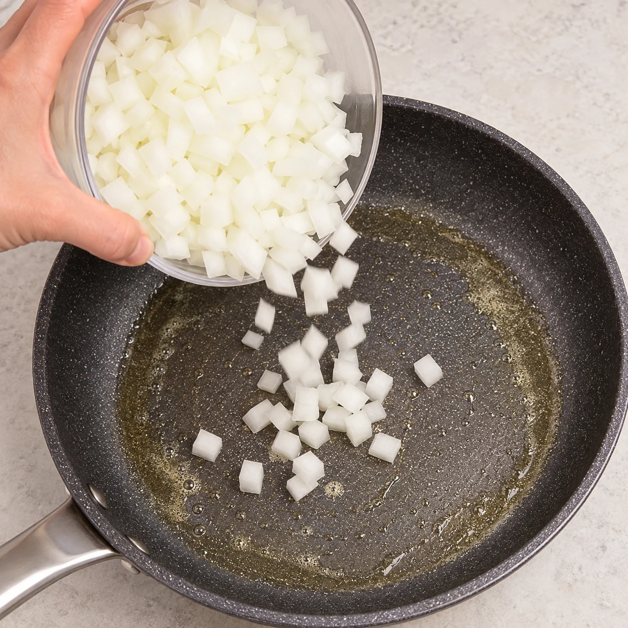 Une main adulte anonyme verse les petits morceaux d’oignons crus depuis un bol neutre dans la poêle contenant une fine pellicule de margarine fondue. Aucun ail, aucune câpre ni épinard encore ajouté.
