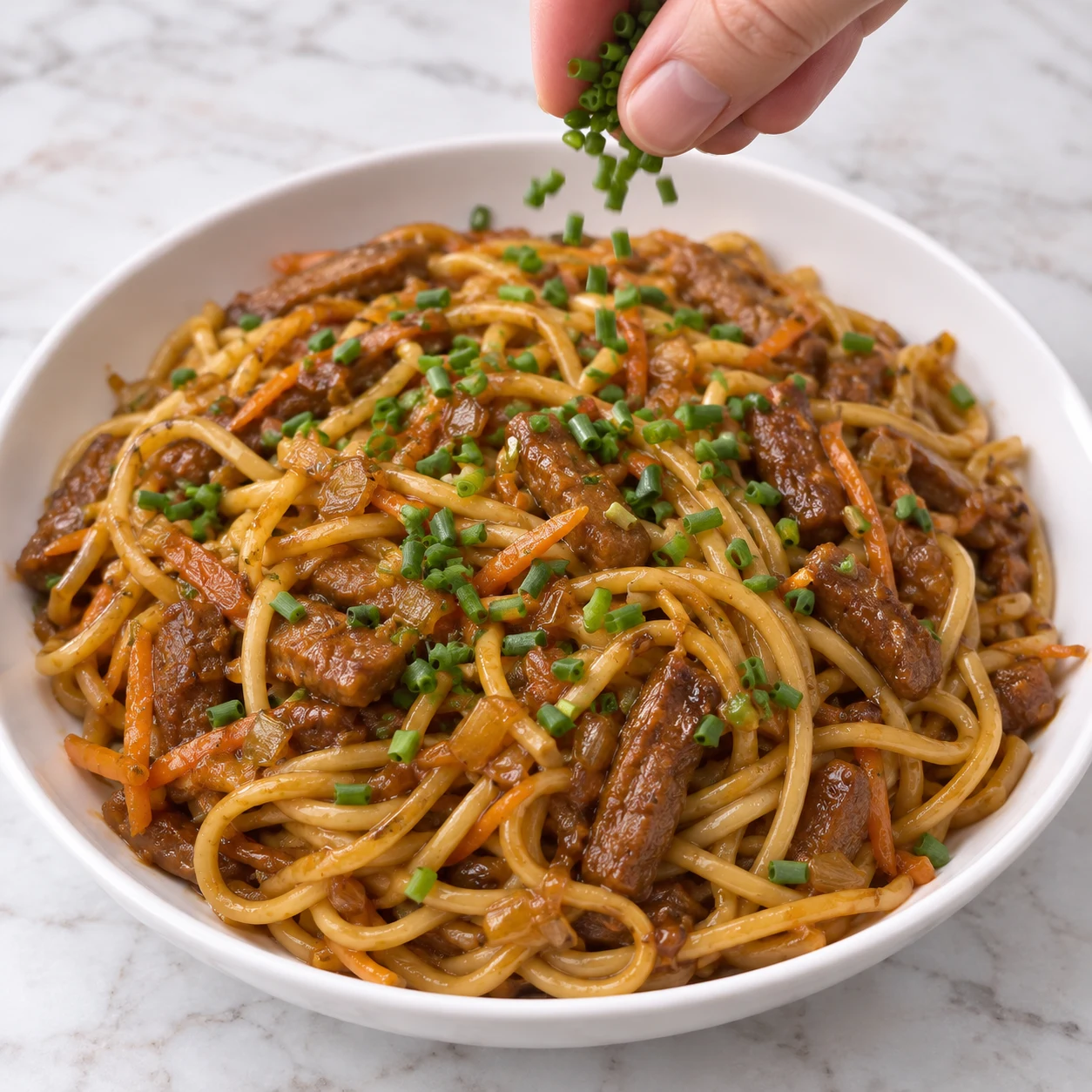 Main anonyme parsemant les nouilles teriyaki, carottes et lamelles végétales de ciboulette ciselée dans une assiette blanche.