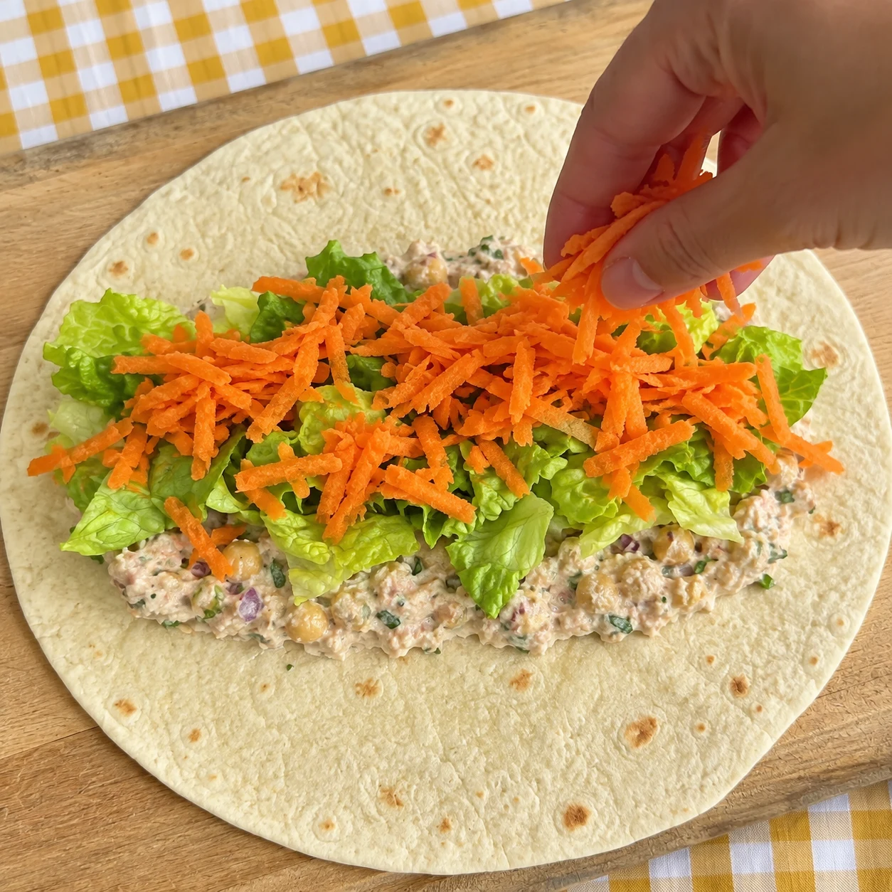 Une main anonyme dépose la carotte râpée orange sur la salade au-dessus de la garniture aux pois chiches dans la tortilla ouverte, pas encore d’avocat.