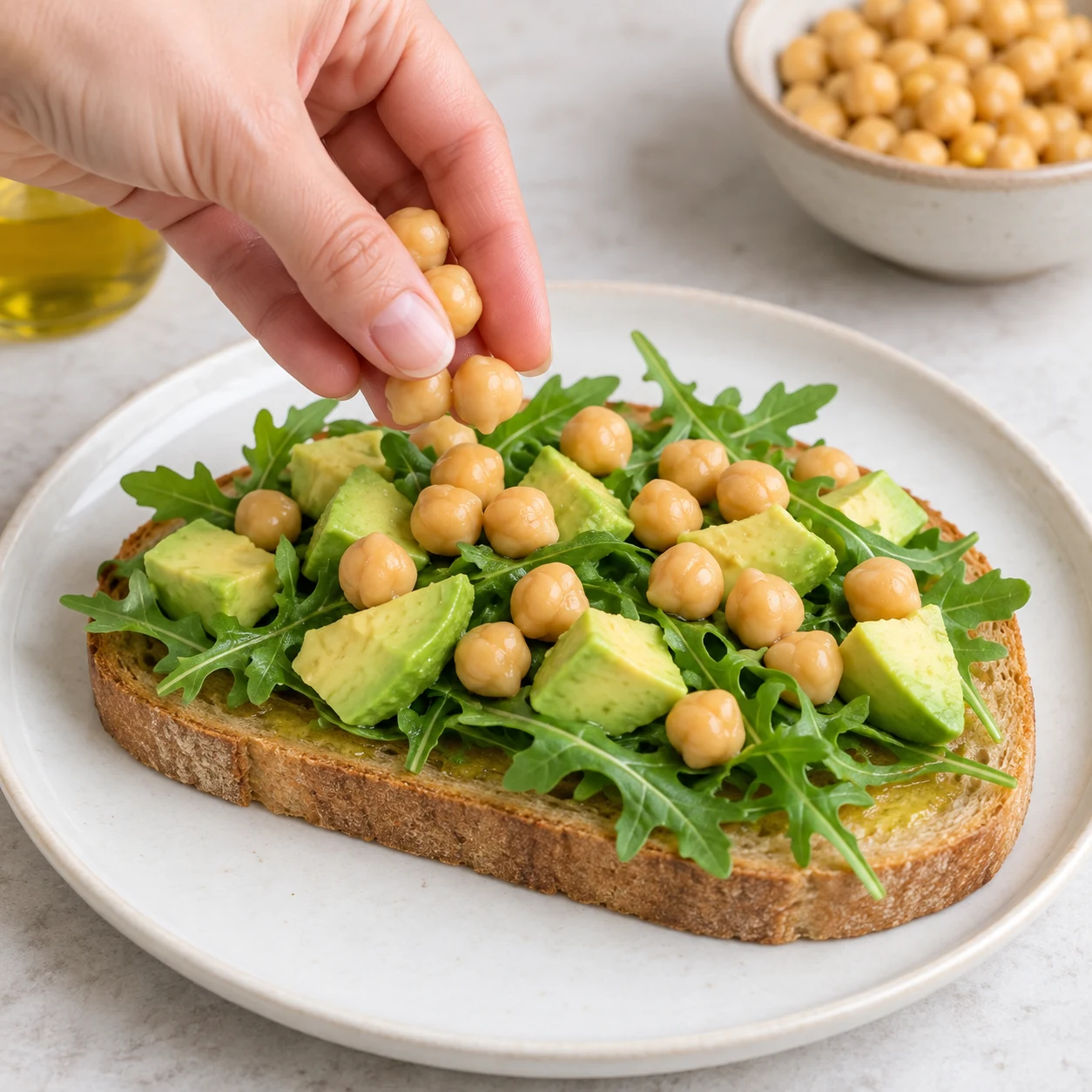Répartis les 100 g de pois chiches égouttés sur la tartine à l’avocat. Photographie carrée de cuisine, ingrédients végétaux finaux seulement, mains adultes anonymes si nécessaires, aucun texte ni visage.