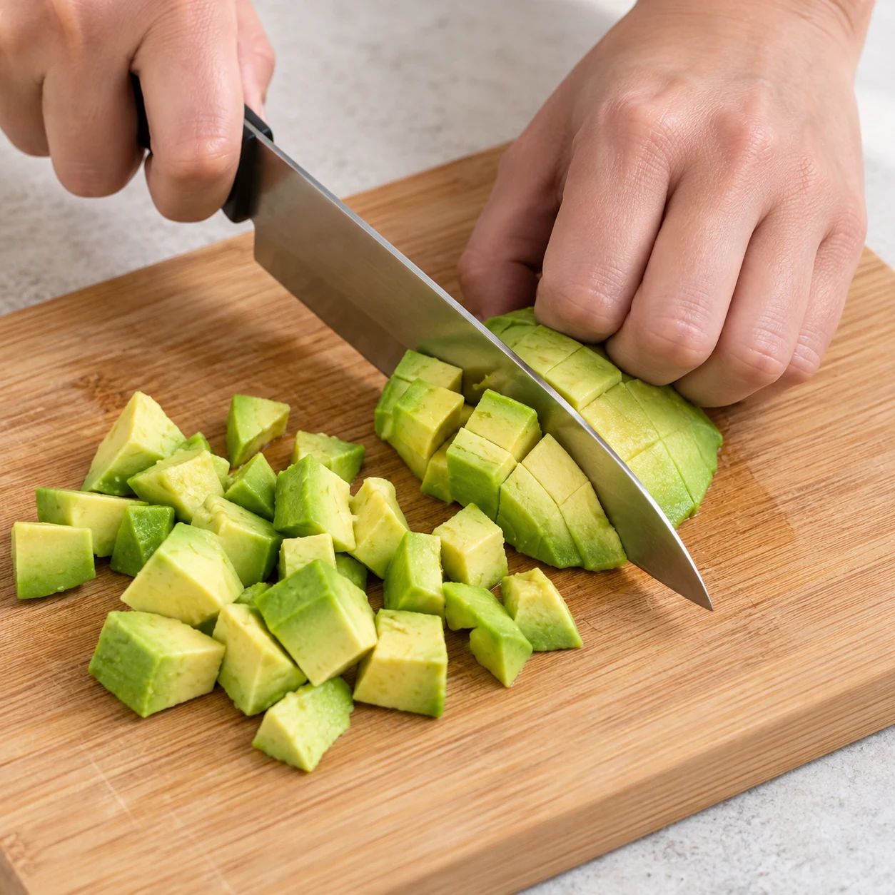 Sur une planche stable, coupe la chair du demi-avocat en petits morceaux avec un couteau, les doigts repliés à distance de la lame. Photographie carrée de cuisine, ingrédients végétaux finaux seulement, mains adultes anonymes si nécessaires, aucun texte ni visage.