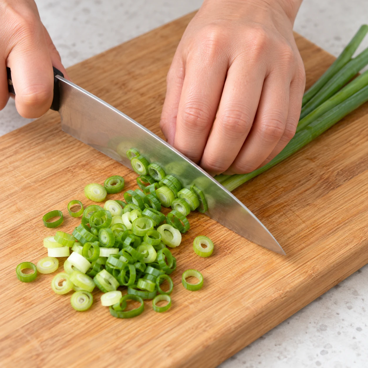Une main adulte anonyme en prise en griffe émince une cébette verte fraîche en fines rondelles sur une planche stable ; aucun champignon ni autre aliment.