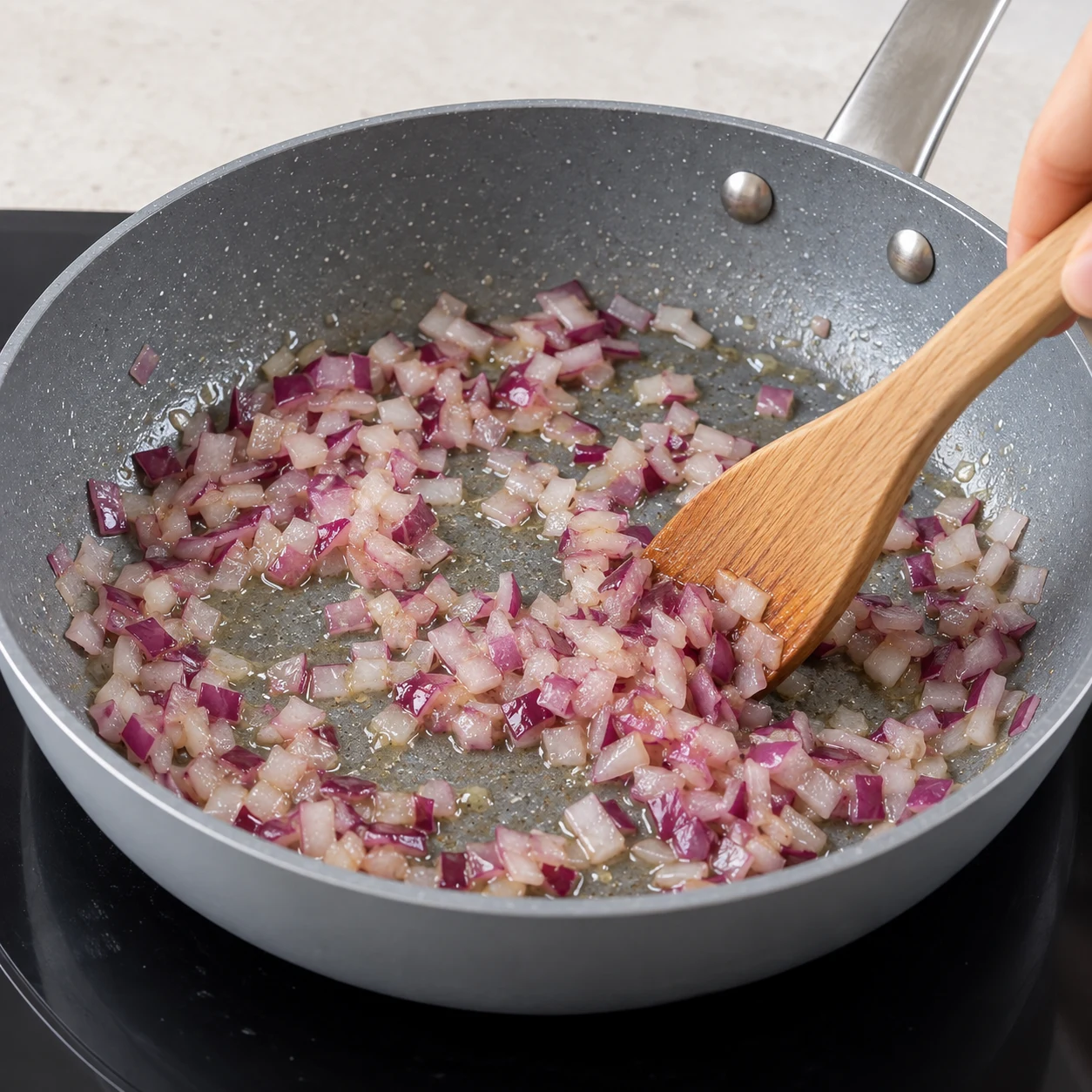 Fais revenir les morceaux d’oignon dans une sauteuse avec un peu d’huile d’olive ; remue pour éviter qu’ils attachent. Montrer seulement les ingrédients de cette phase ; mains anonymes, sauteuse grise et plan de travail pierre pâle.