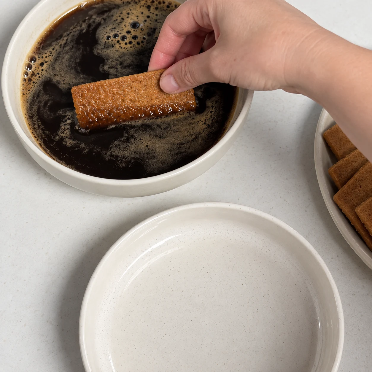 Main adulte trempant un spéculoos brun doré dans un bol de café noir, plat rond vide à côté.