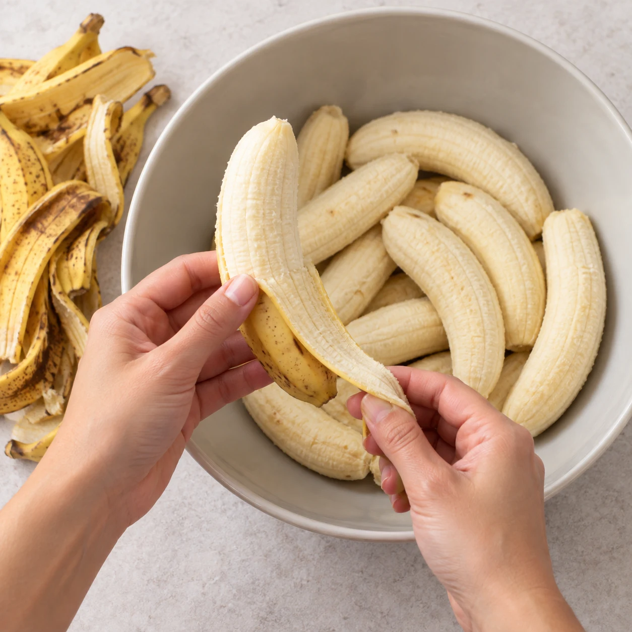 Une seule paire de mains adultes anonymes retire la peau d’une banane mûre au-dessus d’un bol neutre avec des bananes pelées, aucun autre ingrédient.