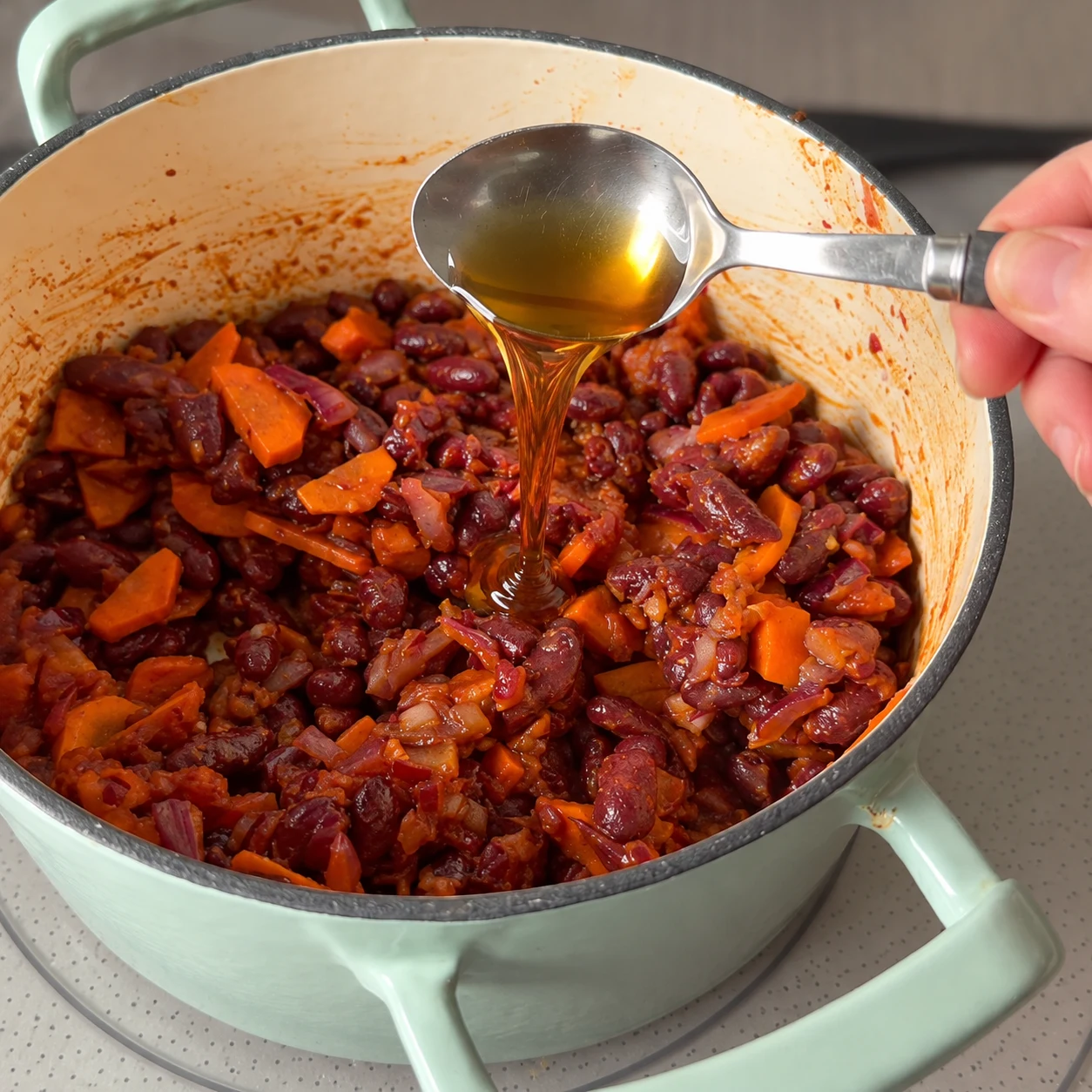 Une photographie carrée : Verse la cuillère à soupe de sirop d’érable sur les haricots. Résultat : Le sirop d’érable est versé dans la cocotte.. Aucun ingrédient futur. Le Cayenne remplace le piment fumé original ; pas de piment fumé ajouté.