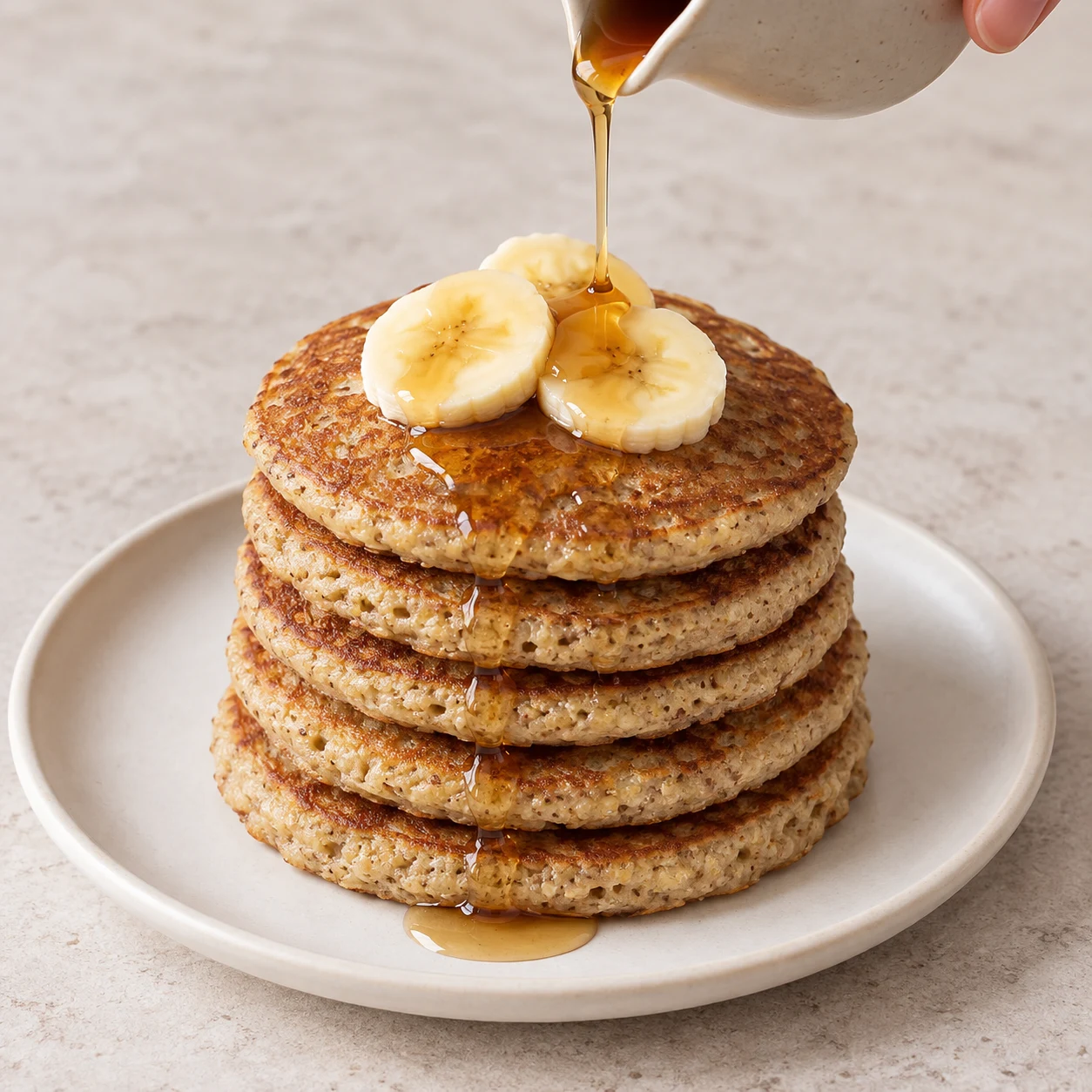 Pile de 6 pancakes dorés mouchetés avec quelques rondelles de banane et sirop, assiette neutre.