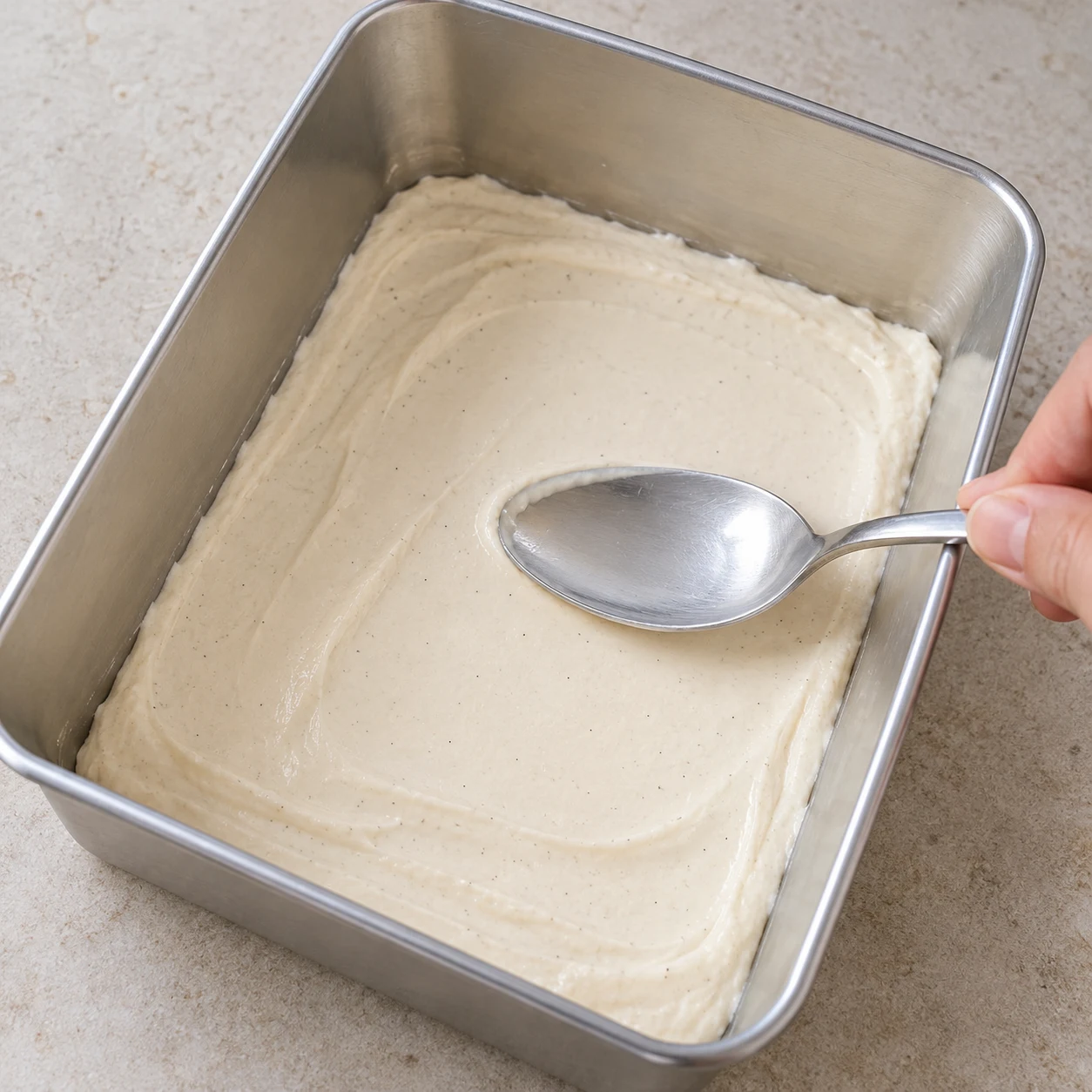 Moule rectangulaire en inox dont le fond est couvert d'une couche lisse de crème beige clair, dos de cuillère qui l'égalise.